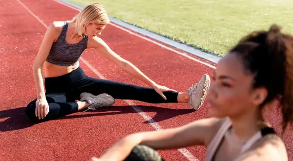 Deux femmes font un exercice de mobilité sur une piste d'athlétisme. L'une est assise, jambe tendue, l'autre est floue au premier plan.