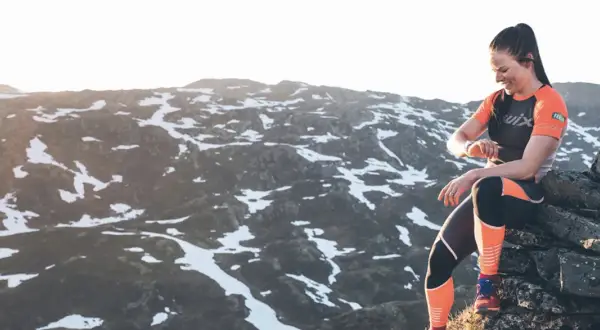 Femme souriante, en tenue de sport, assise sur des rochers en montagne, vérifiant sa montre. Préparation ultra trail dans un paysage enneigé.