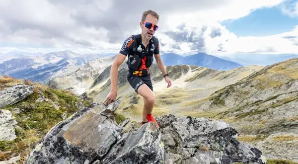Un traileur en plein effort sur un sentier rocailleux, le regard déterminé, incarne l'ultra spirit dans un paysage montagneux spectaculaire.