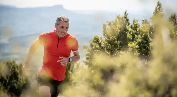 Un homme en tenue de sport orange et noire, souriant, prêt à commencer la course à pied sur un sentier de montagne.