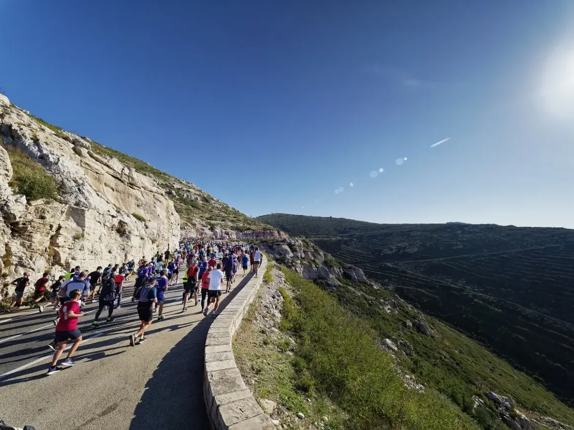 Course de running sur une route côtière, avec un grand groupe de coureurs affrontant le dénivelé Marseille Cassis sous un ciel bleu éclatant.