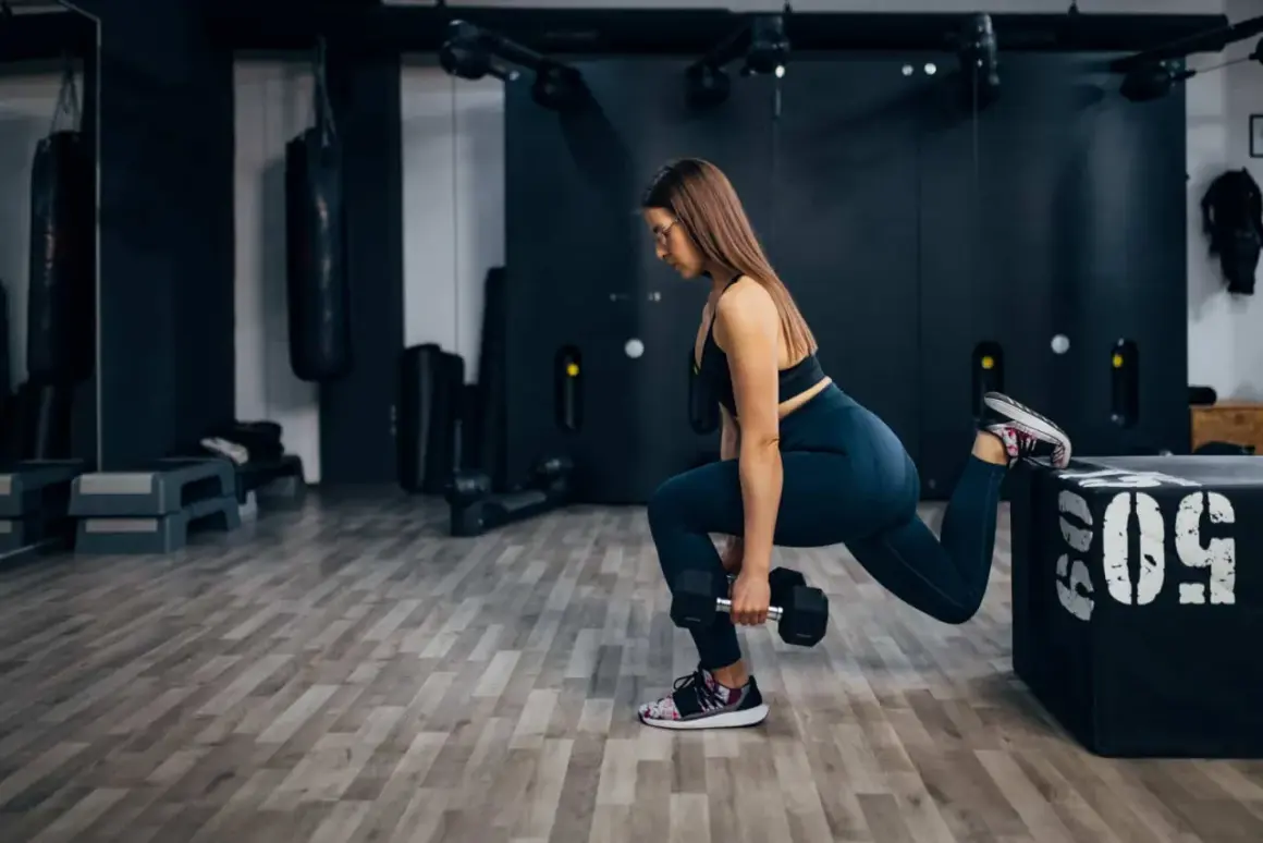 Femme effectuant une fente bulgare avec haltère, entraînement de jambes intense dans une salle de sport moderne.