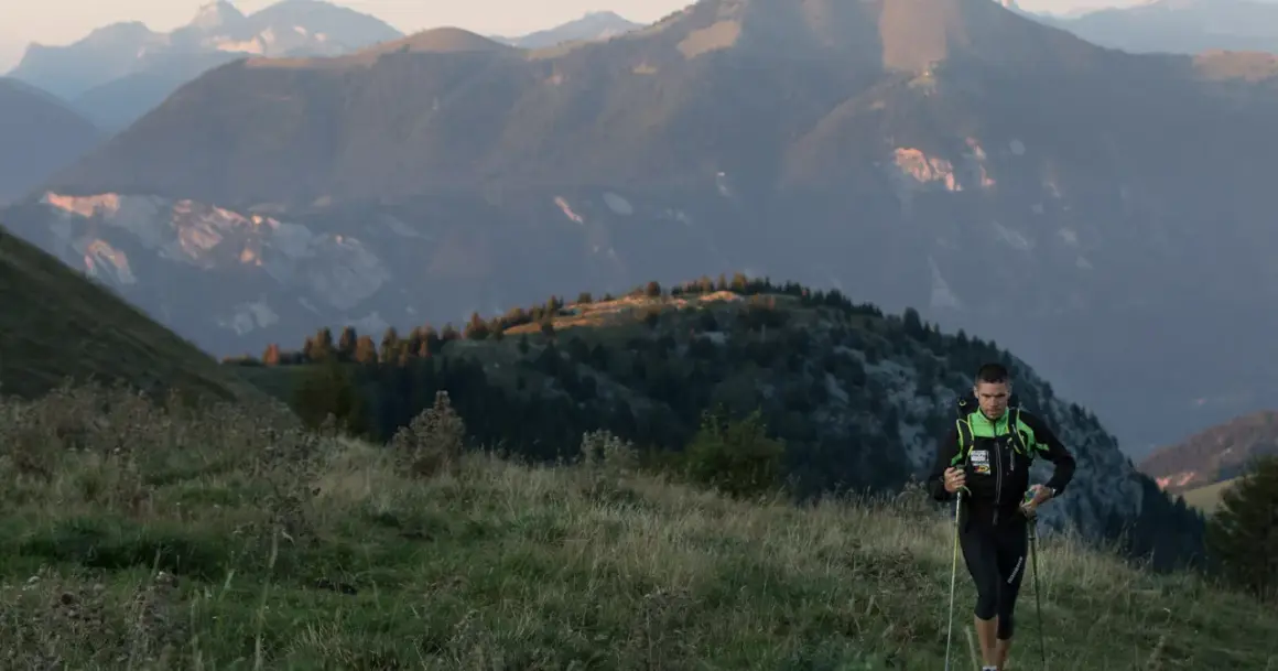 Un coureur de trail avec des bâtons monte une colline herbeuse, avec des montagnes en arrière-plan.
