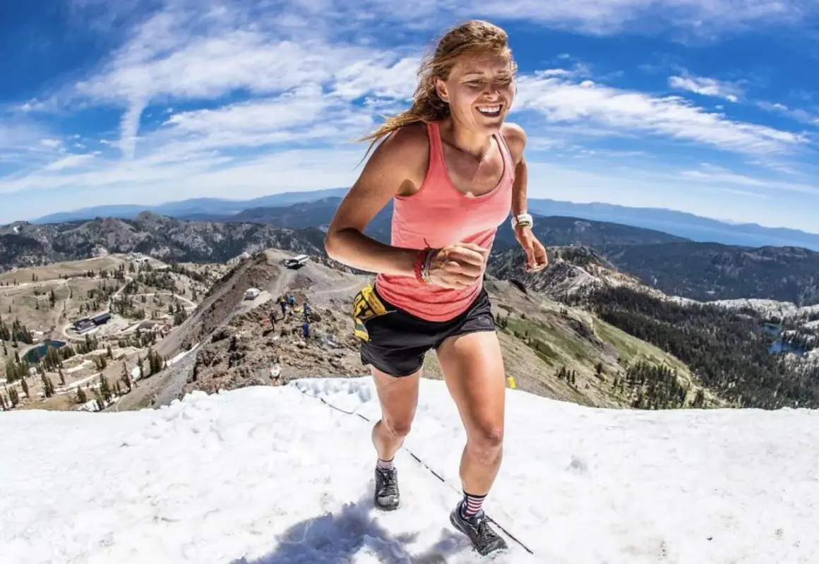Une coureuse en plein effort, le visage rayonnant, dévale une pente enneigée en montagne. Son **endurance active** est mise à l'épreuve dans ce paysage spectaculaire.