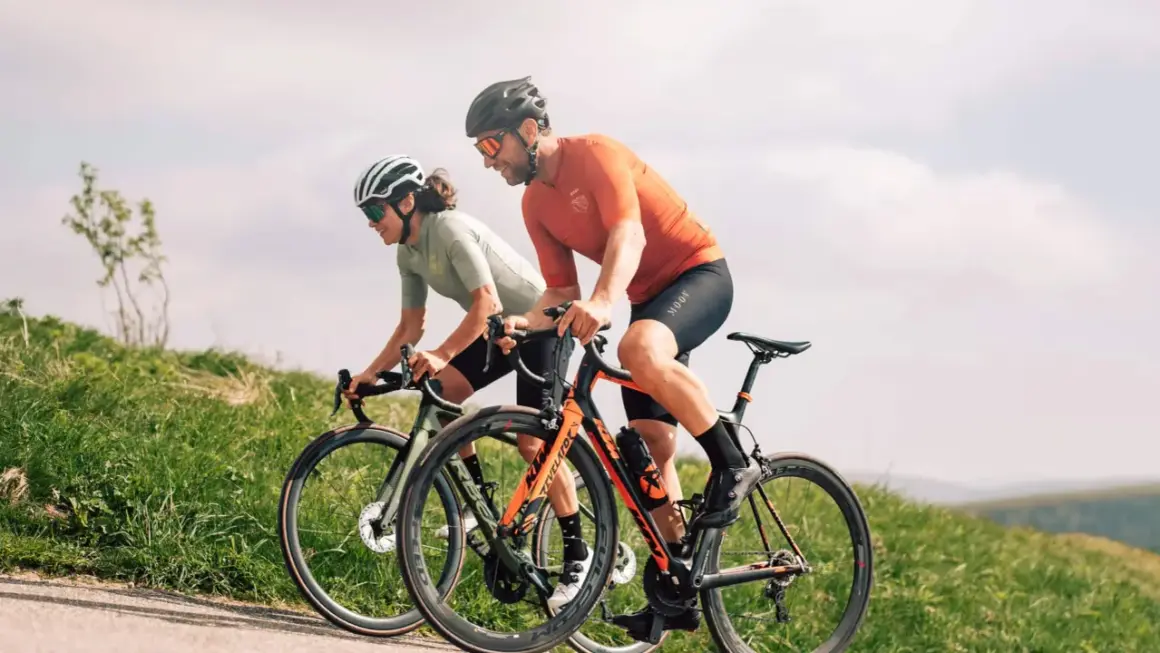 Deux cyclistes en plein **entrainement croisé** sur une route de campagne, profitant du paysage verdoyant sous un ciel nuageux.