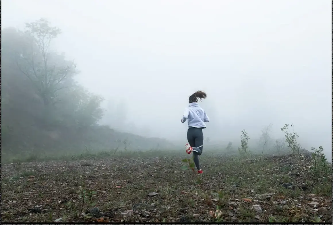 Femme courant dans le brouillard, une scène de préparation physique trail. Le paysage est brumeux, avec des arbres et un terrain rocailleux.