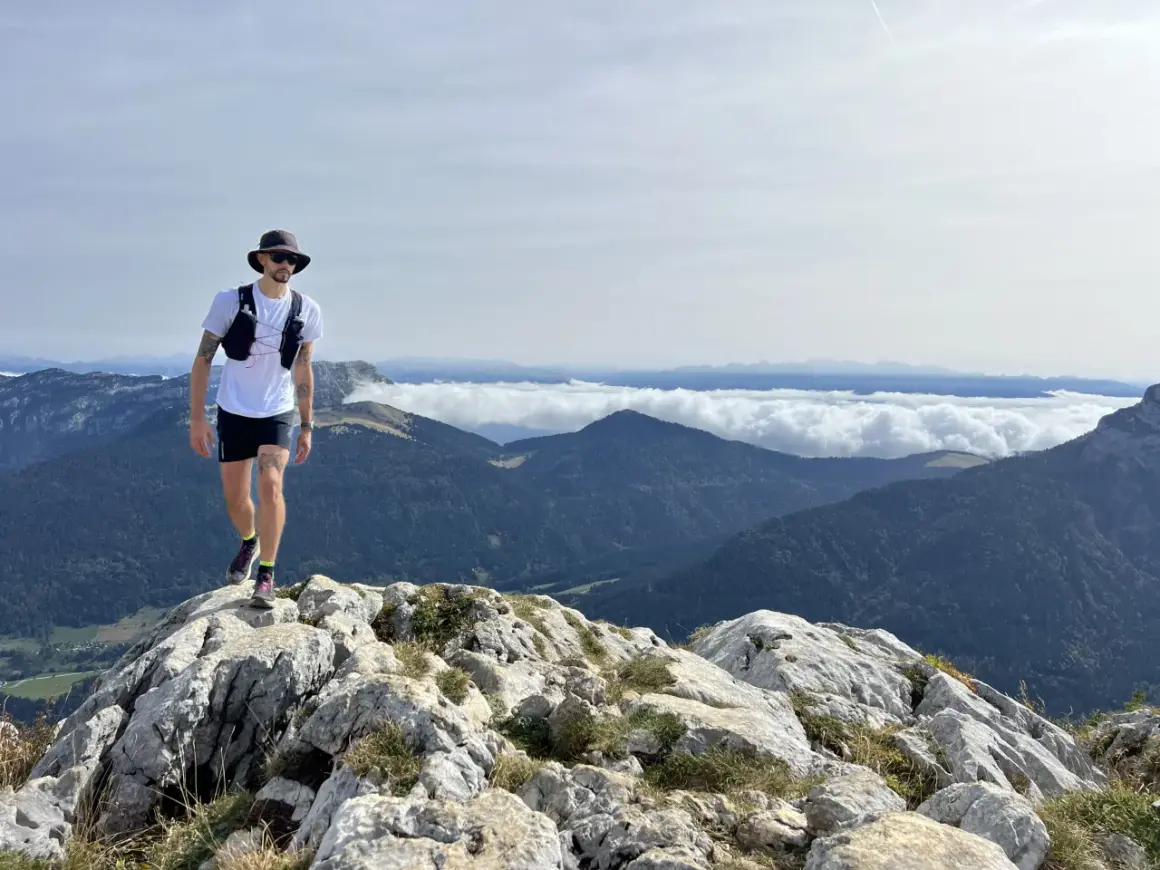 Un homme en tenue de trail, chapeau et lunettes, marche sur un sentier rocheux avec une vue imprenable sur les montagnes. Un week-end choc trail inoubliable.
