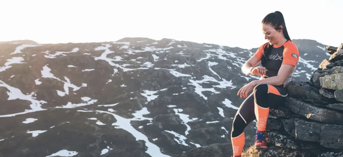 Femme souriante, en tenue de sport, assise sur des rochers en montagne, vérifiant sa montre. Préparation ultra trail dans un paysage enneigé.