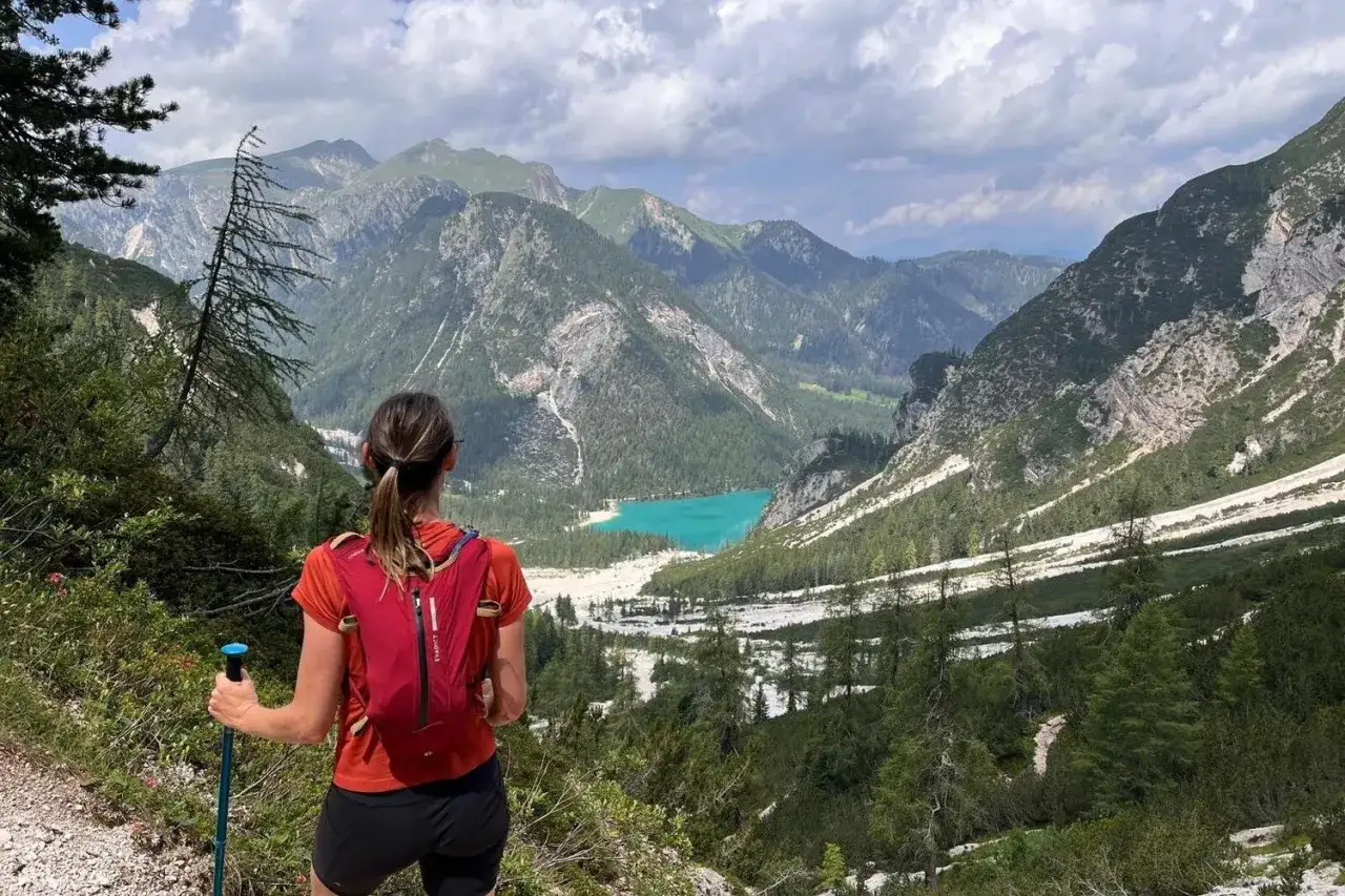 Vue panoramique sur un lac turquoise entour&eacute; de montagnes verdoyantes. Une femme en tenue de sport, sac &agrave; dos rouge et b&acirc;tons de marche, profite du paysage lors d'un entrainement trail 20 km.
