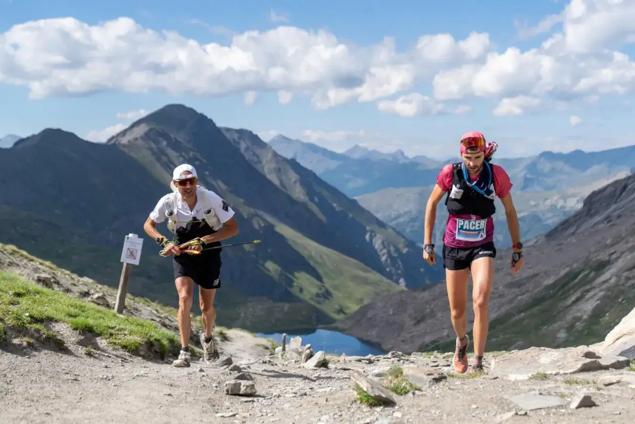 Deux coureurs s'élancent sur un sentier de montagne escarpé, un lac bleu scintillant en contrebas. L'un d'eux porte un dossard