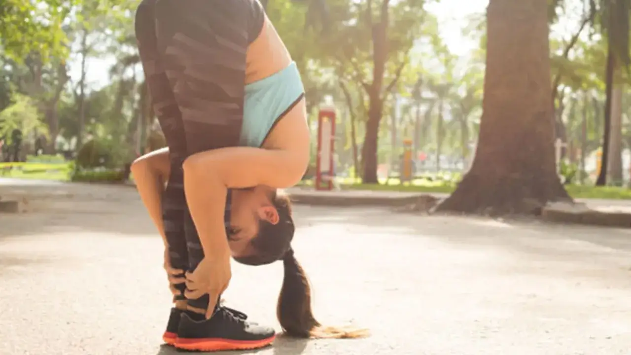 Femme en tenue de sport effectuant un &eacute;tirement abdos en se penchant en avant, mains agrippant ses chevilles, dans un parc ensoleill&eacute;.