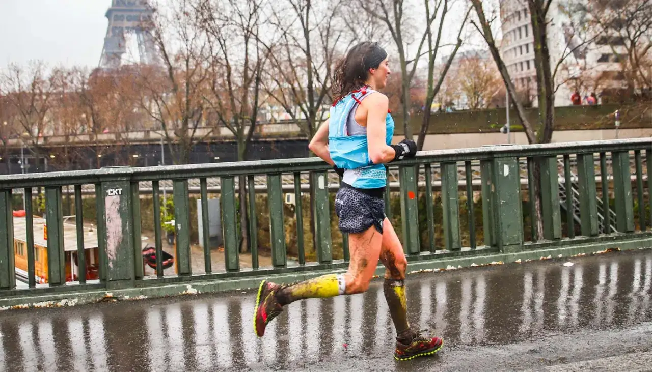 Une coureuse boueuse traverse un pont &agrave; Paris, la Tour Eiffel en arri&egrave;re-plan. Elle court sous la pluie, ses jambes couvertes de terre.