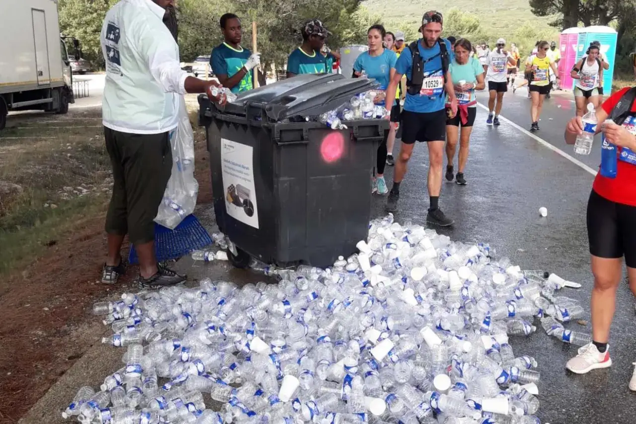 Des coureurs passent devant une benne à ordures débordant de bouteilles d'eau. Un défi pour savoir où courir à Marseille sans laisser de trace.