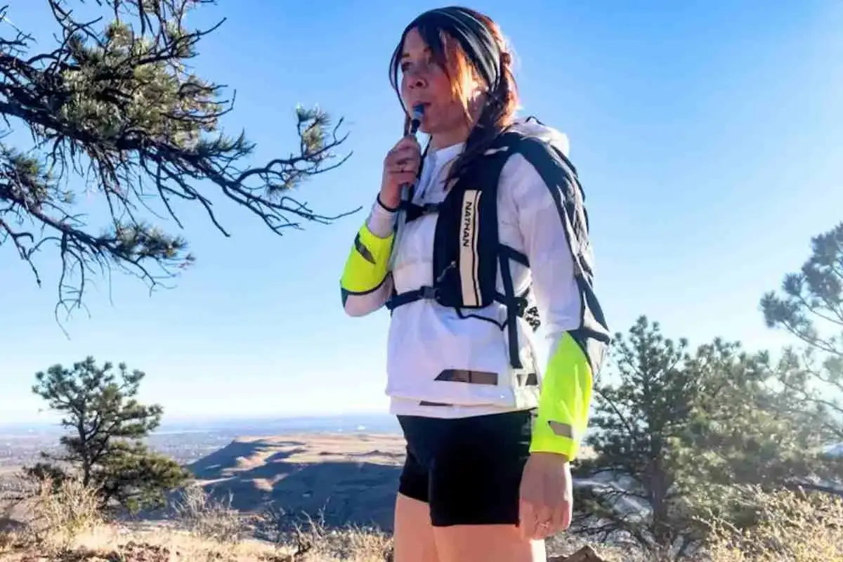 Femme buvant dans une poche à eau, prête pour la préparation ultra trail. Veste de course Nathan, bras réfléchissants, sur fond de ciel bleu et de nature.