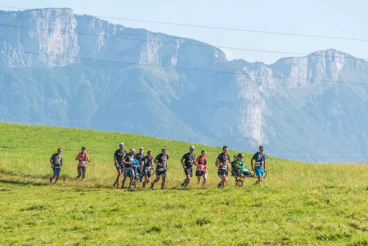 Une équipe court solidaire lors d'une joelette course, traversant une prairie verdoyante avec des montagnes majestueuses en arrière-plan.