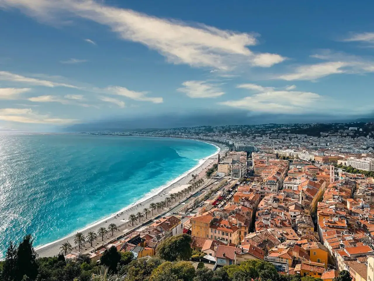 Vue panoramique de la Baie des Anges &agrave; Nice, avec sa plage de galets, ses palmiers et la ville s'&eacute;tendant &agrave; perte de vue. Id&eacute;al pour une promenade ou pour courir &agrave; Nice.