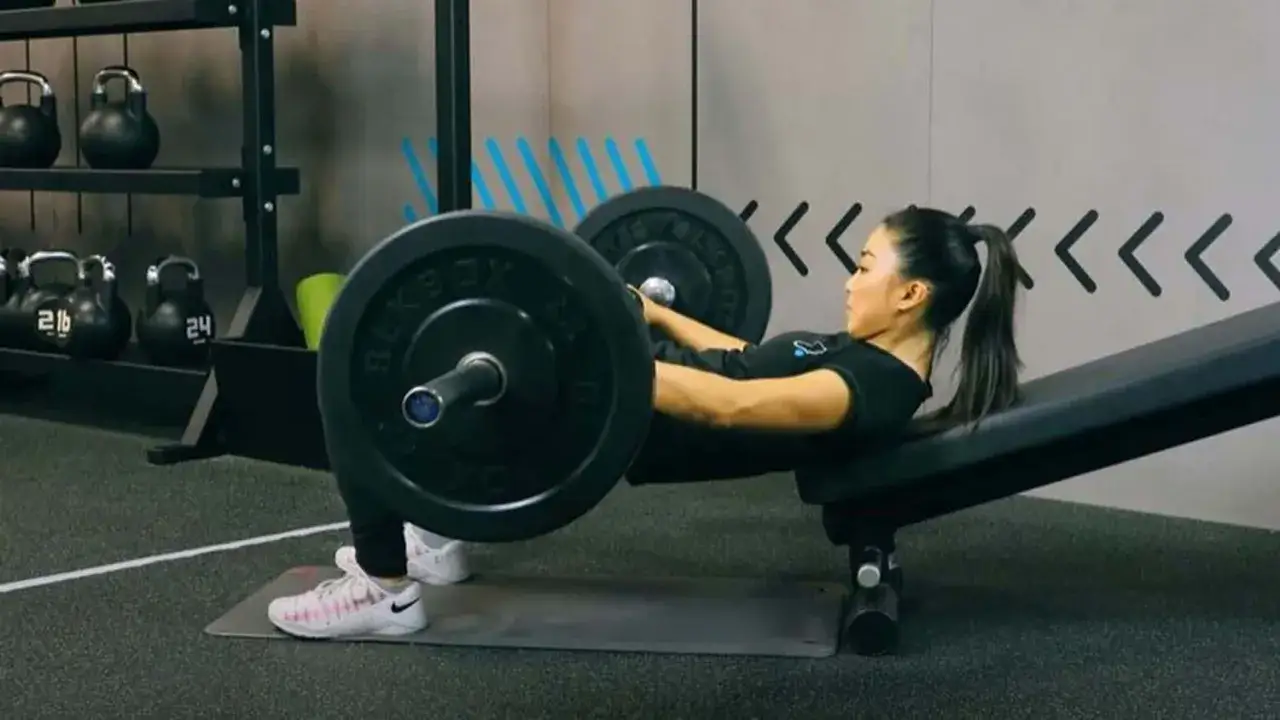 Femme effectuant un hip thrust avec une barre sur un banc incliné dans une salle de sport.