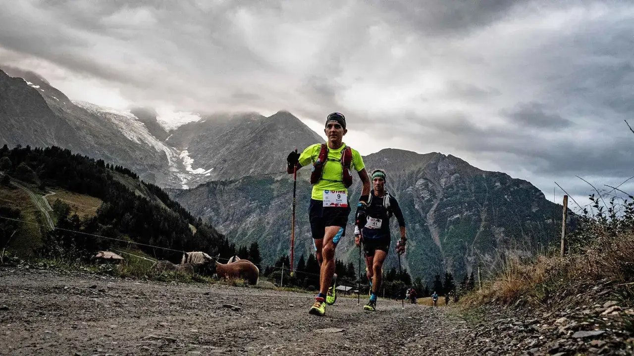 Deux coureurs en pleine pr&eacute;paration mentale trail, gravissent un sentier rocailleux en montagne. Le ciel est couvert, les sommets enneig&eacute;s en arri&egrave;re-plan.