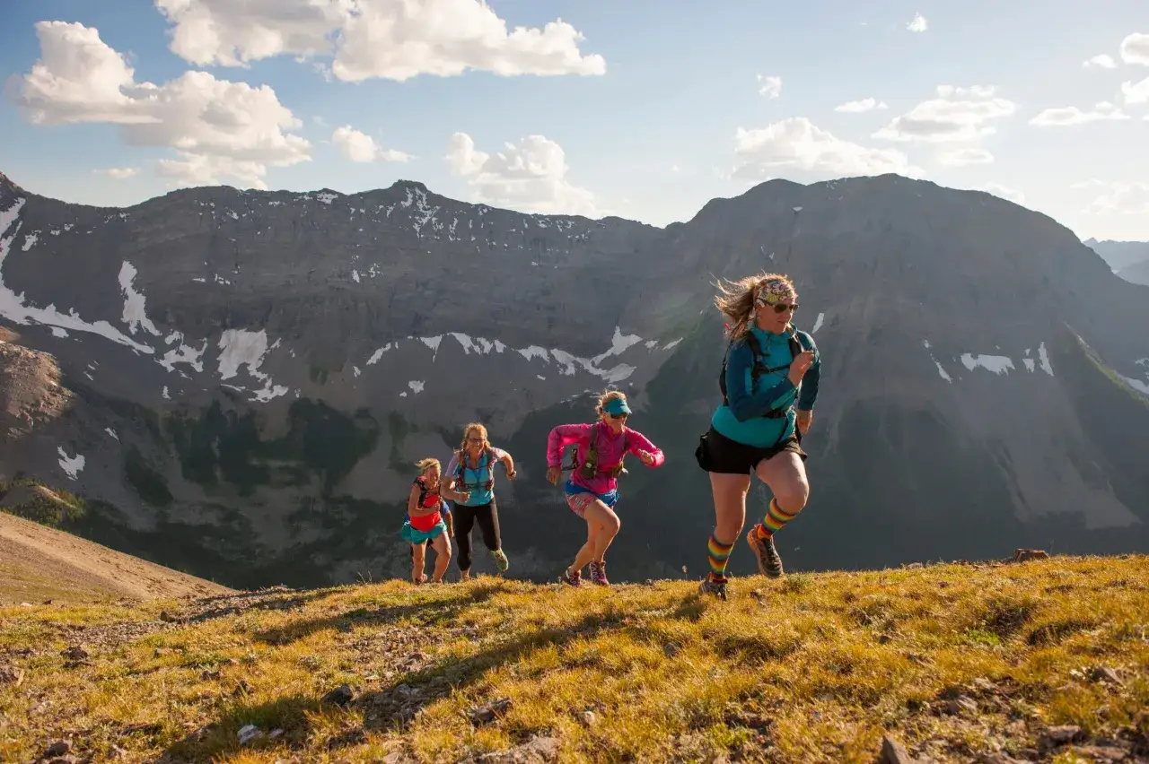 Quatre femmes courent sur un flanc de montagne herbeux, profitant du meilleur trail sous un ciel bleu et nuageux.