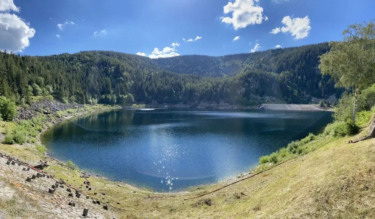 Lac paisible entour&eacute; de for&ecirc;ts denses, parfait pour une randonn&eacute;e sur un trail vosgien. Le soleil scintille sur l'eau bleue.