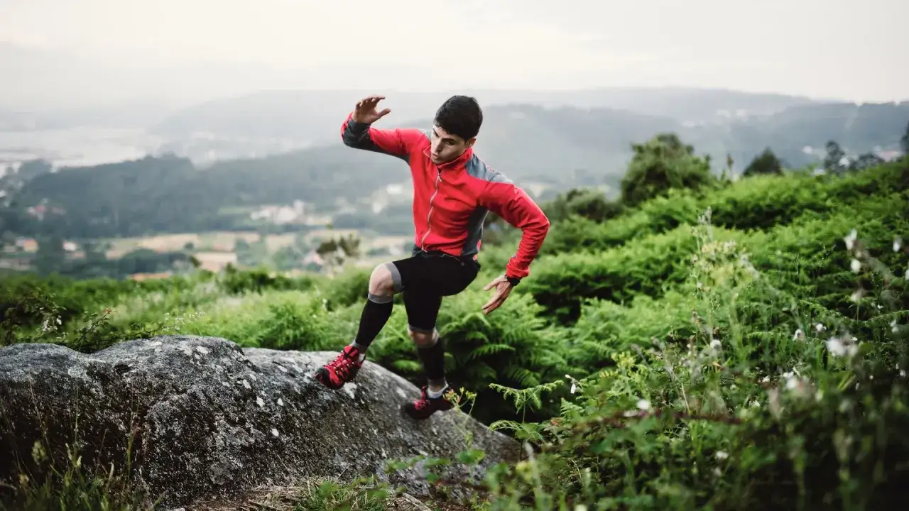 Un homme en tenue de sport rouge et noire fait du trail run sur un rocher, entour&eacute; de v&eacute;g&eacute;tation luxuriante et d'une vue sur la vall&eacute;e.