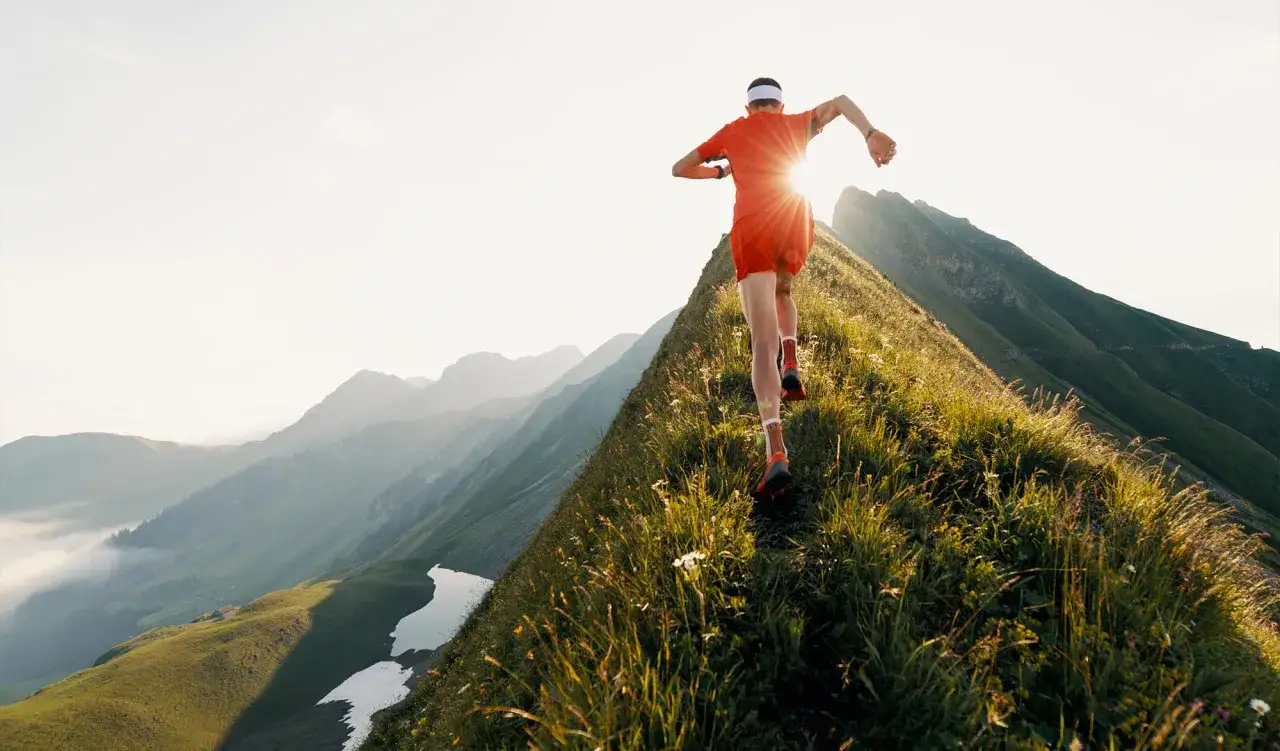 Un coureur en tenue orange dévale une crête herbeuse, le soleil levant illuminant le paysage montagneux. Une démonstration d'endurance active.