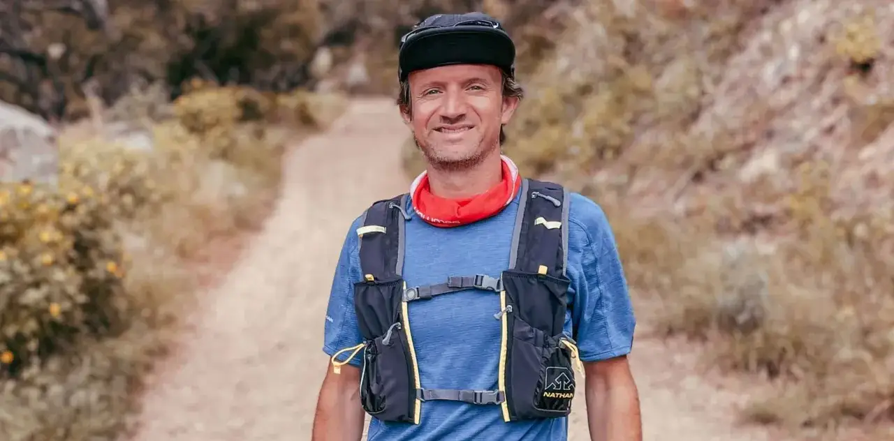 Un homme souriant, prêt pour une trail longue distance, porte un sac de trail et un bandana rouge. Le chemin s'étend derrière lui.