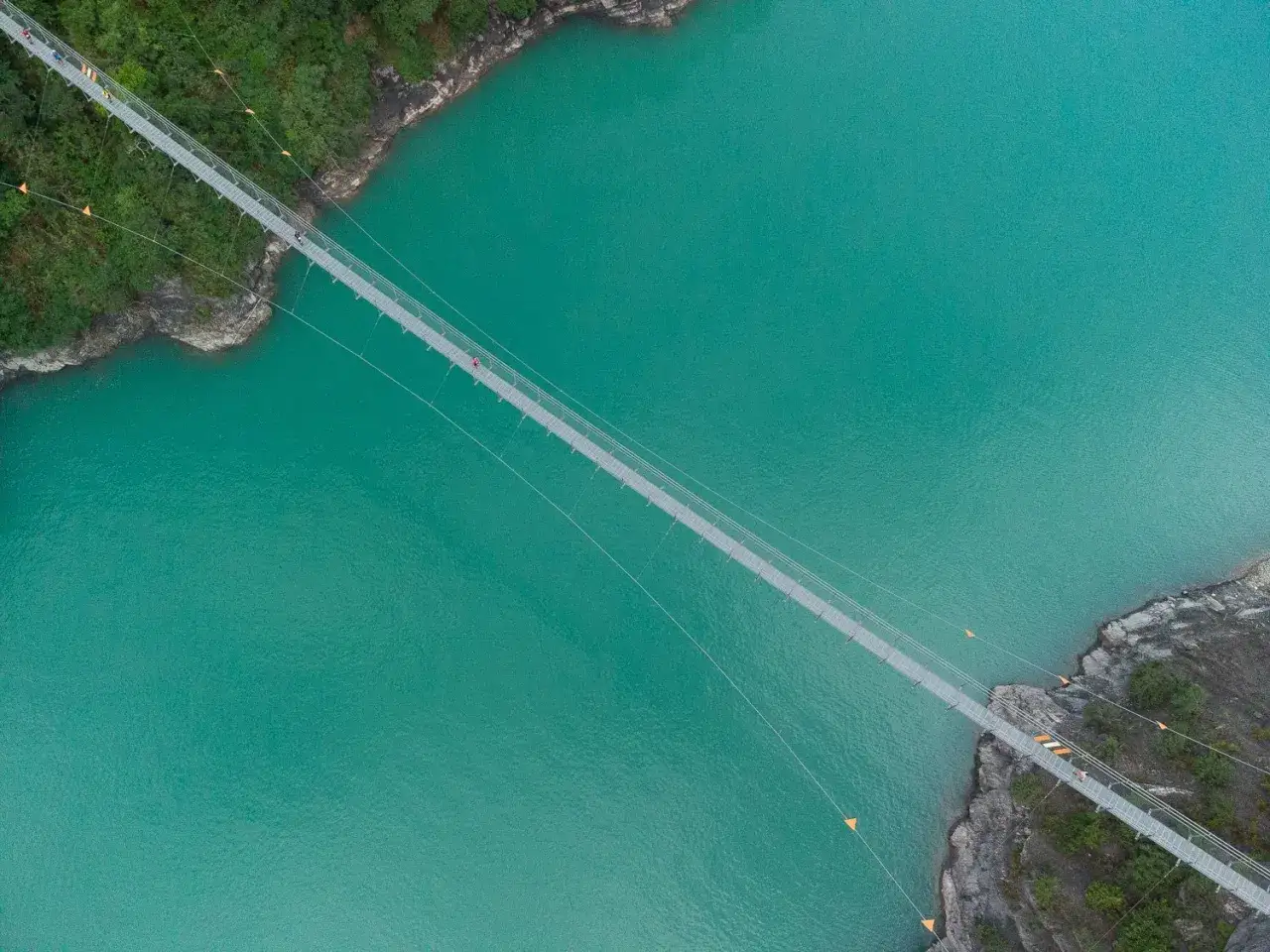 Vue aérienne d'un pont suspendu traversant une eau turquoise, parfait pour le maratrail des passerelles. Des coureurs s'aventurent sur la structure.