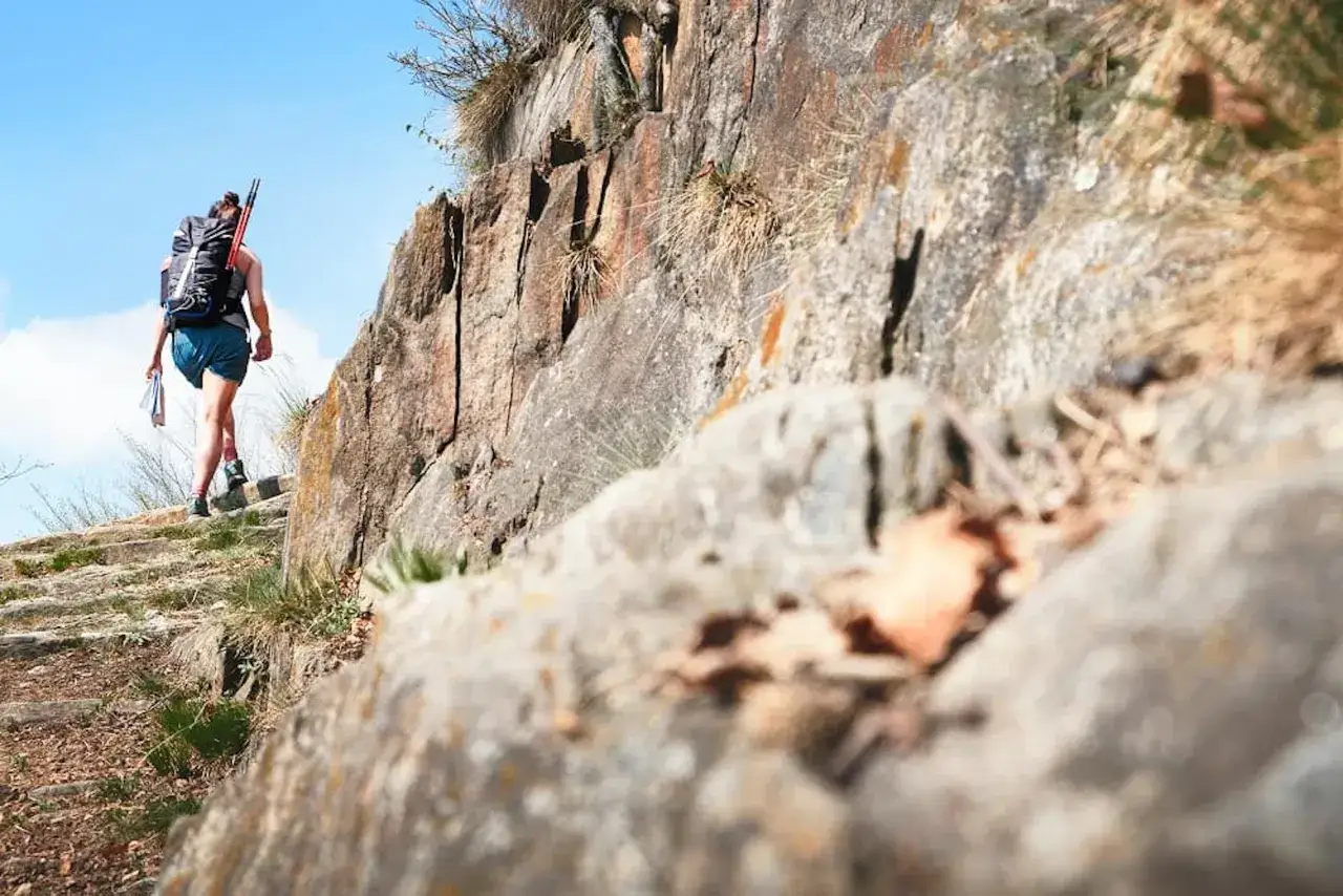 Une randonneuse avec un grand sac à dos monte un sentier montagneux rocailleux sous un ciel bleu clair.