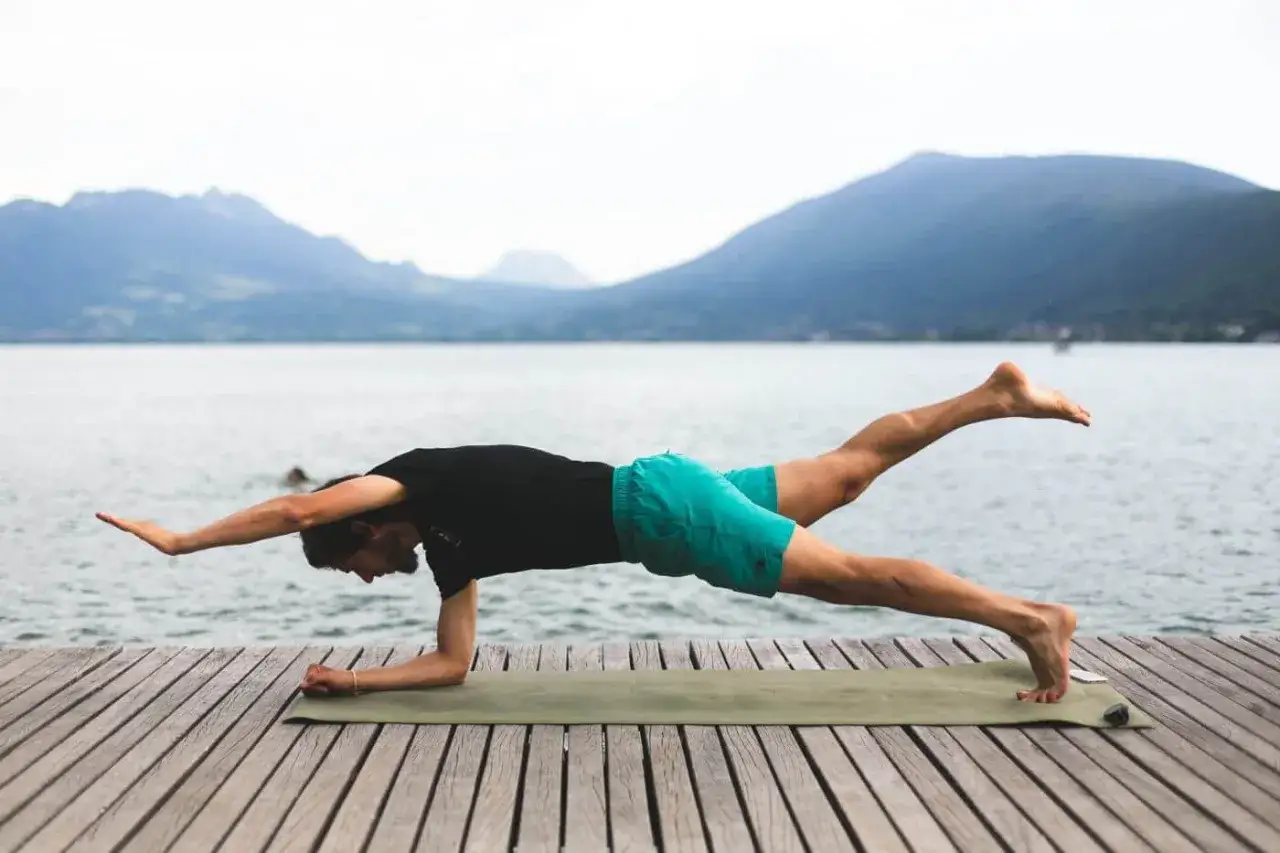 Un homme réalise un exercice oblique sur un tapis, bras et jambe levés, avec un lac et des montagnes en arrière-plan.