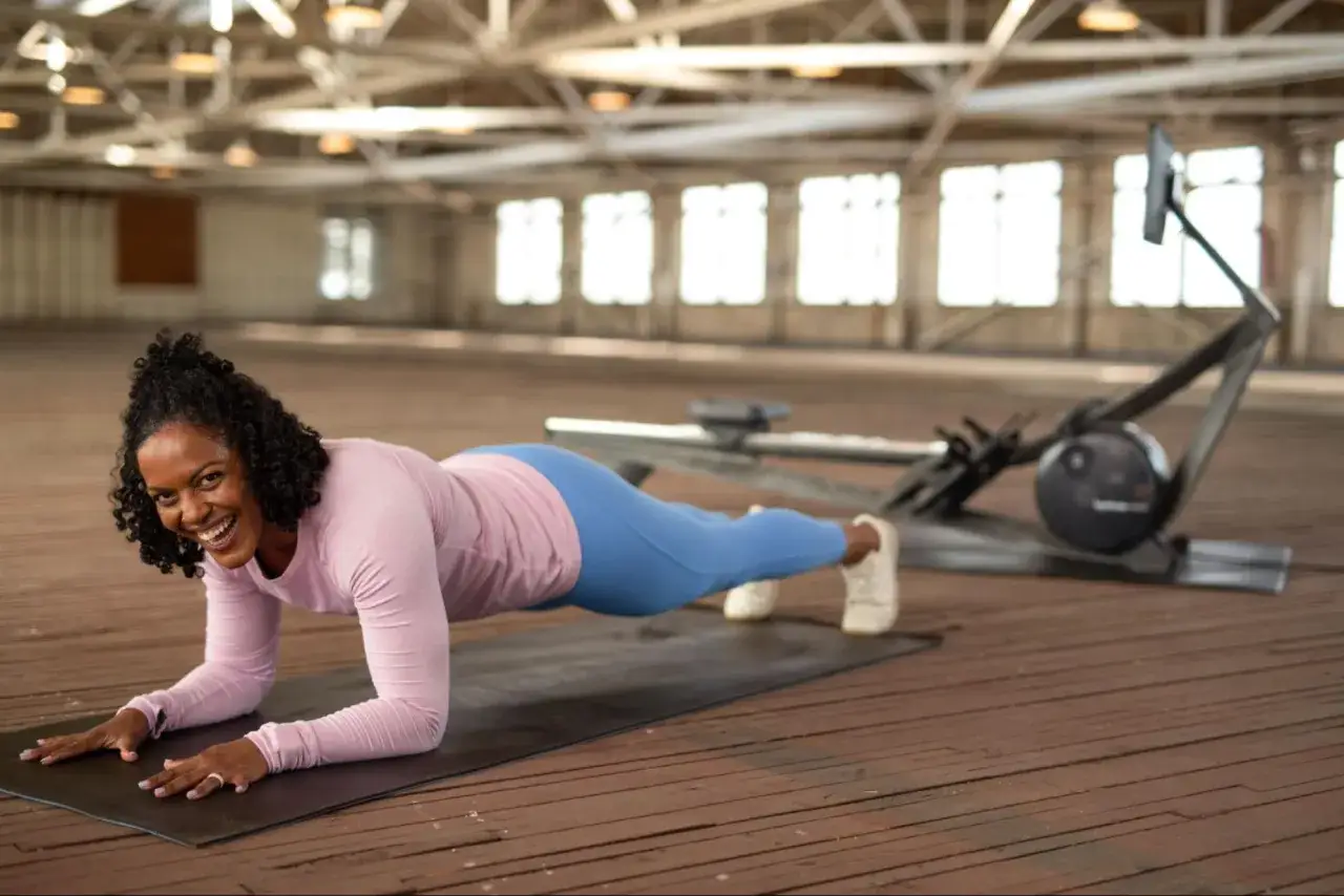 Femme souriante en plein gainage ventre sur un tapis, avec un rameur en arrière-plan.