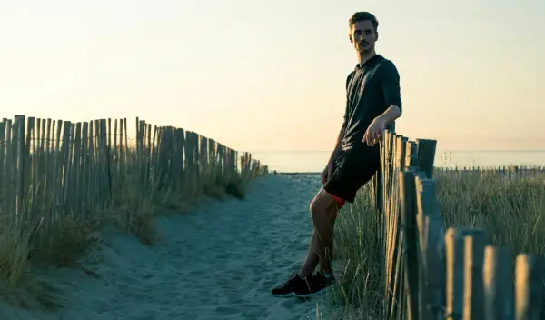 Homme sportif, en tenue de course, se repose contre une clôture de bois sur une plage au coucher du soleil. Il a atteint son poids de forme.