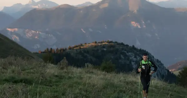 Un coureur de trail avec des bâtons monte une colline herbeuse, avec des montagnes en arrière-plan.