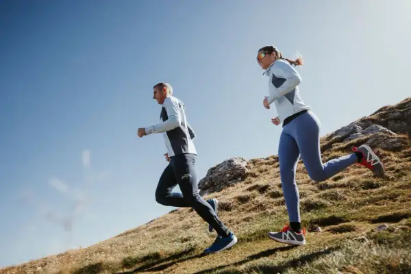 Couple en pleine préparation ultra trail 100 km, courant sur un sentier de montagne rocailleux sous un ciel bleu.