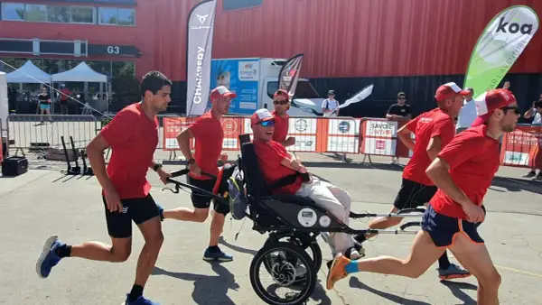 Une équipe court avec un homme en fauteuil roulant lors d'une course en joëlette. Ils portent tous des t-shirts rouges et des casquettes.