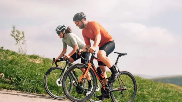 Deux cyclistes en plein **entrainement croisé** sur une route de campagne, profitant du paysage verdoyant sous un ciel nuageux.