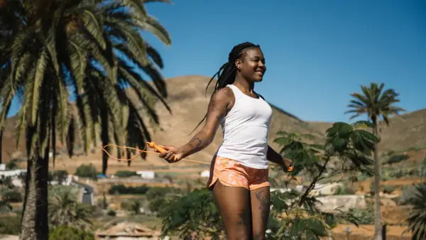 Femme souriante faisant de l'exercice corde à sauter sous un ciel bleu, entourée de palmiers et de collines.