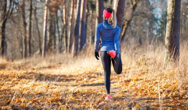 Une coureuse fait un étirement après trail dans une forêt automnale, entourée de feuilles mortes et d'arbres.