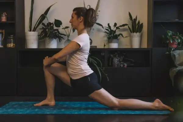 Femme effectuant un étirement TFL sur un tapis de yoga bleu, entourée de plantes vertes.