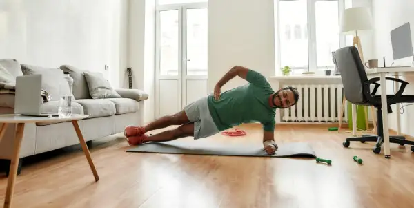 Un homme souriant pratique le gainage latéral sur un tapis de yoga dans son salon.