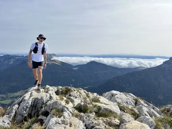 Un homme en tenue de trail, chapeau et lunettes, marche sur un sentier rocheux avec une vue imprenable sur les montagnes. Un week-end choc trail inoubliable.