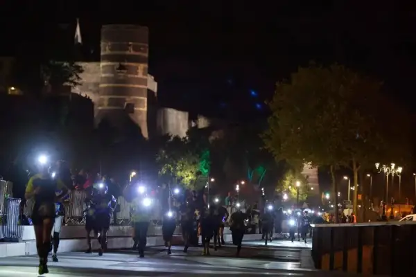 Courseurs illuminés par des lampes frontales, traversant la ville pour le trail de l'apocalypse, avec un château en arrière-plan.