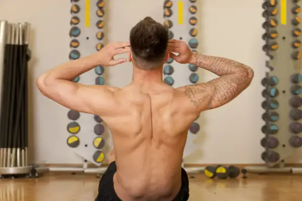 Homme musclé effectuant un exercice pour le dos, mains derrière la tête, dans une salle de sport avec des haltères en arrière-plan.
