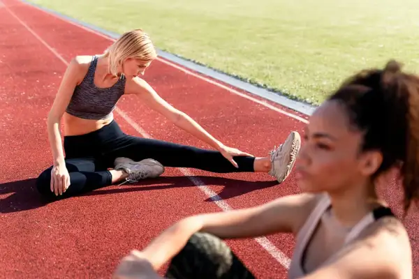 Deux femmes font un exercice de mobilité sur une piste d'athlétisme. L'une est assise, jambe tendue, l'autre est floue au premier plan.