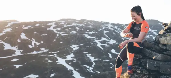 Femme souriante, en tenue de sport, assise sur des rochers en montagne, vérifiant sa montre. Préparation ultra trail dans un paysage enneigé.